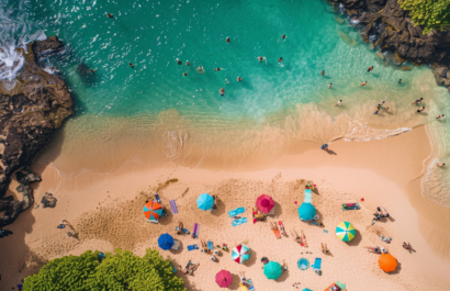 An aerial view of Honomu Beach Park with people sunbathing, kids building sandcastles, and vibrant umbrellas dotting the sandy shore.